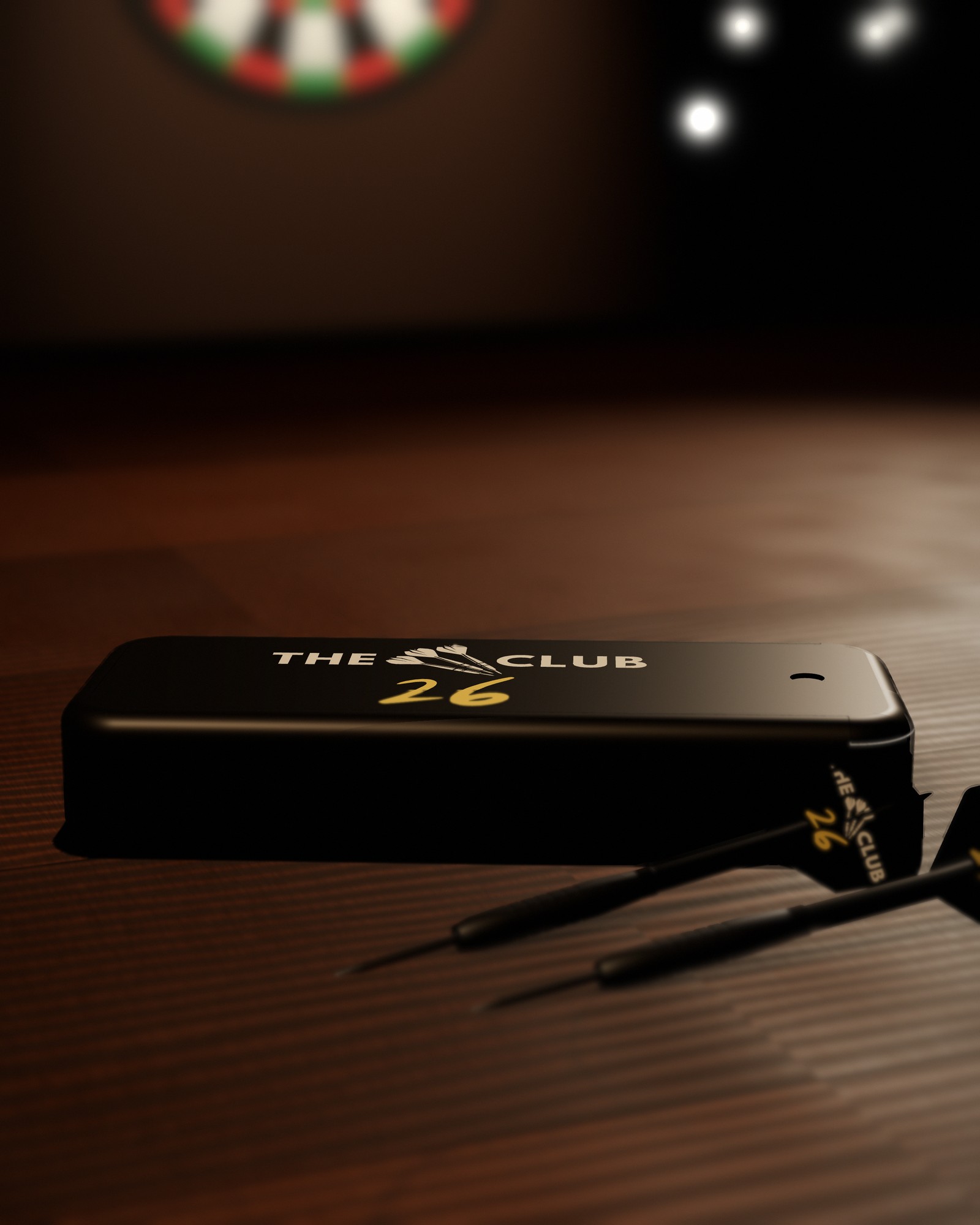 Render of the black slide-top dart case, closed, with The 26 Club logo in white and gold on the lid, on a pub table beside two darts