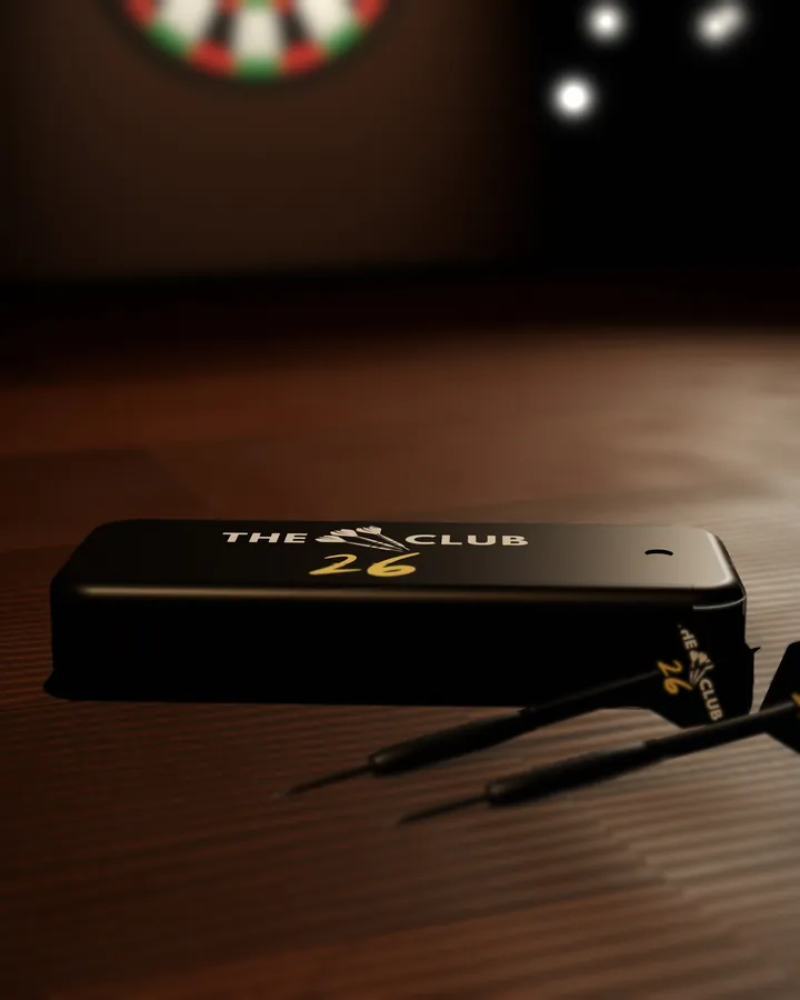 Render of the black slide-top dart case, closed, with The 26 Club logo in white and gold on the lid, on a pub table beside two darts